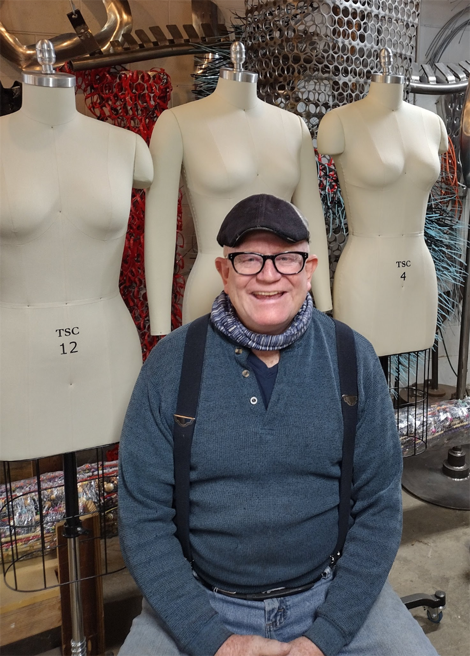 Gerald Fitzpatrick sitting in front of three dress mannequins and various metal materials, wearing a grey cap, glasses, scarf, overalls, and navy sweater