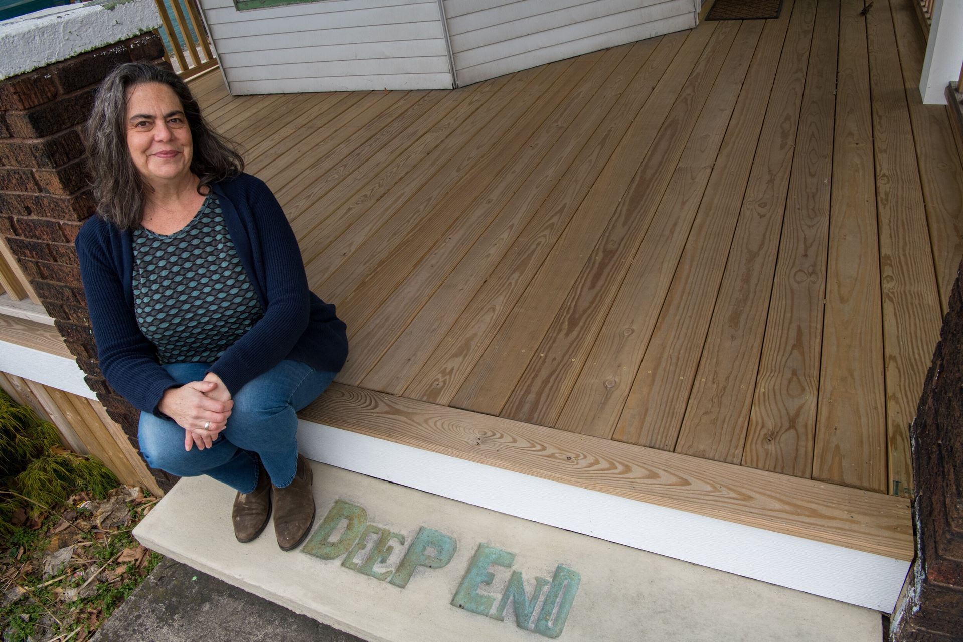 Portrait of Bellamy Printz sitting on a porch with the words "Deep End" at her feet.  She is wearing a green and blue shirt with a blue sweater and jeans. Her hair is dark and wavy.