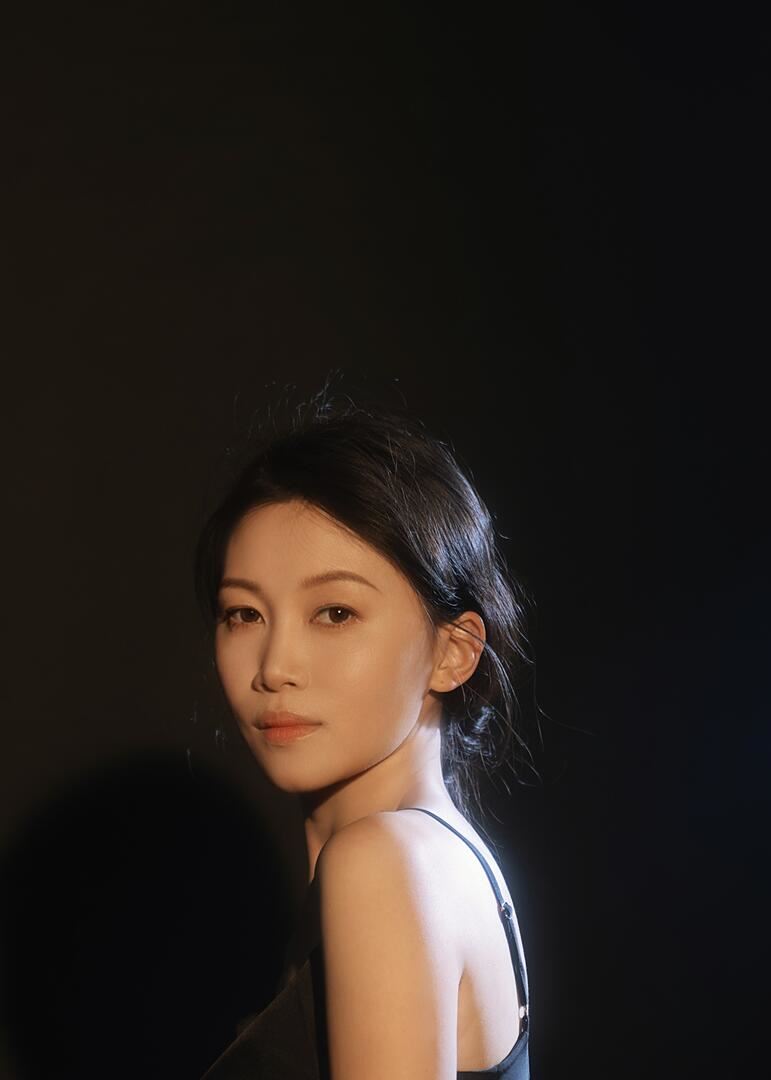 Portrait of Weiting Wei standing in profile and looking out over her shoulder. Background is black.  Wei has long dark hair and is wearing a black tank top.